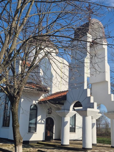 A vertical, full shot shows the Church of the Holy Trinity in Shumentsi, Bulgaria. The church is white with a rounded dome on top. A tree with bare branches is in the foreground, partially obscuring the church. The sky is blue and cloudless. The lower section of the church has windows and a door with an arched top. The church is bordered by a wall of light brown stone blocks.