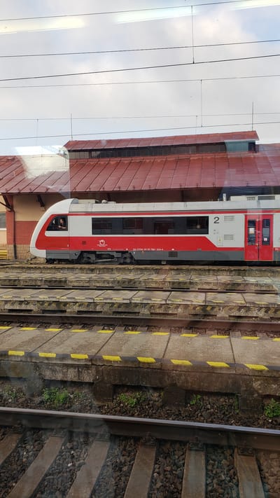 The image shows a red and white train stopped at a station in Margecany, Slovakia. The train is a modern electric multiple unit, indicated by the number "2" on its side. A person is visible in the driver's cabin. The train is on a track with a concrete platform next to it, marked with yellow lines. In the background, a building with a red tiled roof is visible, which is part of the Margecany train station. Overhead power lines for the electric train are also present. The sky is overcast.