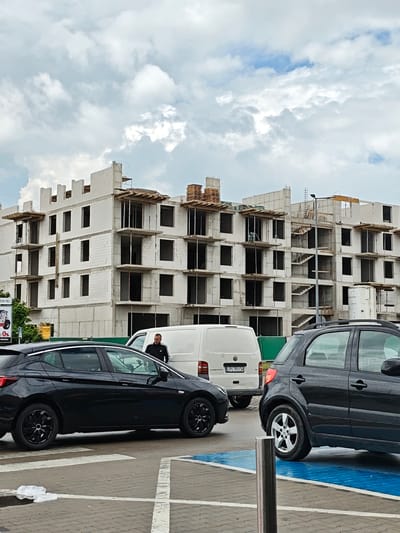 An overcast sky looms over a construction site in Mława, Poland, where a multi-story building is under construction. The building's exterior walls are made of light-colored bricks, with visible concrete balconies and staircases. Wooden scaffolding supports the unfinished upper levels. In the foreground, several cars are parked in a lot. A black hatchback with a distinctive wheel design is on the left, and a dark-colored SUV is on the right. A white van with the license plate "LPU 95134" is parked between the cars and the construction site. A man in a dark tracksuit stands near the van, facing the construction site. The parking lot is paved with gray cobblestones, marked with white lines and blue painted sections. Debris, possibly plastic, is scattered on the ground near the black hatchback. A street sign with partially obscured text and a "0%" indicator is visible behind the construction site on the left. Trees and greenery line the background.