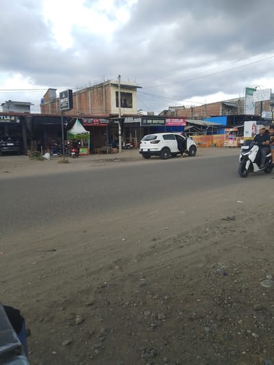 This media file captures a street scene in Gampongpaya, Indonesia, under a cloudy sky, suggesting it is daytime. The foreground shows a dusty, unpaved area bordering a paved road. Several buildings line the road, appearing to be shops and businesses.
Prominently featured are two vehicles: a white SUV with its door open and a person partially visible inside, and a scooter with two people riding on it, moving along the road. The scooter riders appear casually dressed.
Various signs are visible on the buildings. One sign reads "AMRISTYLE," another "WM BOUTIQUE WM DIMSUM," and another advertises "ES TELER CREAM DURIAN." There's also a sign for "OPPO" and a billboard with text that includes "12 16 YANG MELEGEND." These signs suggest a commercial area with diverse offerings, including fashion, food, and electronics.
A green and white tent structure is set up in front of one of the businesses, possibly for outdoor sales or seating. A motorcycle is parked near the tent. The overall scene depicts a bustling, everyday commercial street with pedestrian and vehicular activity. The weather appears to be overcast, with a grey, cloudy sky.