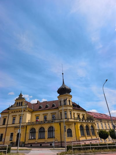 The image shows a bright yellow building with a red-tiled roof and a prominent black dome tower, located in the city of Vysoké Mýto, Czechia. The building features classical architectural elements, including arched windows, decorative cornices, and multiple stories. A flag, appearing to be the flag of the Czech Republic, is visible on a balcony. The sky above is a clear blue with wispy white clouds. In the foreground, there is a street with a pedestrian crossing, a lamppost, and some greenery. The scene appears to be a daytime view of a public building within an urban setting.