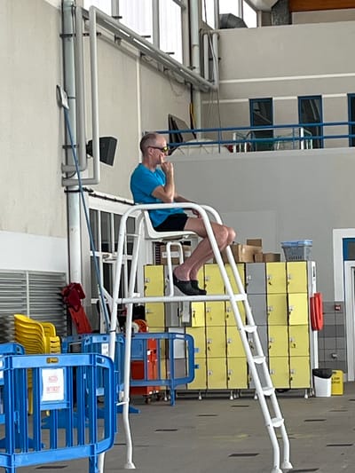 A man is seen on the side of a pool. The man is wearing black slip-on shoes, black shorts, and a short-sleeved blue shirt. The scene is located in Andrézieux-Bouthéon, France. Behind the man are white metal bars, yellow and gray lockers, and blue and yellow plastic chairs. A long, white horizontal pole runs behind the man as well.