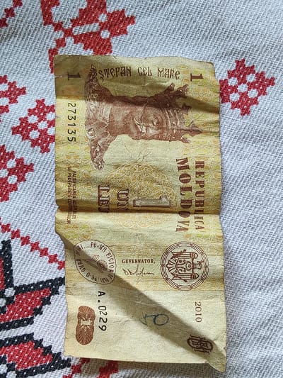 The image shows a wrinkled one-leu banknote from Moldova, featuring a portrait of Stephen the Great. The banknote is placed on a white fabric with a red embroidered traditional pattern. The banknote has a serial number "273135" and "A.0229", and the year "2010". The location of Ucrainca, Moldova, is not visible in the image.