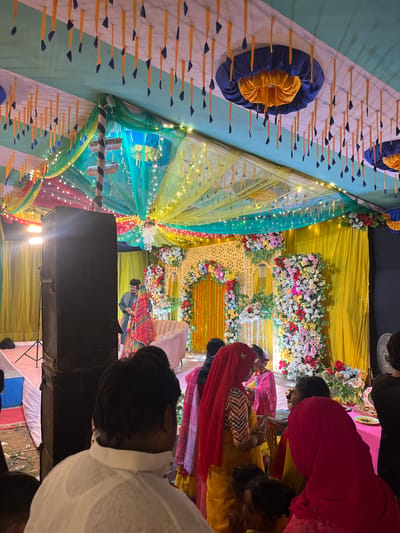 The image depicts an indoor event, likely a wedding ceremony, in Savar, Bangladesh, characterized by elaborate decorations within a large covered space or tent.
**Environment:** The setting is an enclosed area covered by a multi-colored canopy made of fabric drapes in green, yellow, blue, and red. The ceiling is adorned with numerous yellow fabric strips, each tipped with a small dark blue element, and decorative blue and yellow circular fabric features. Numerous string lights are suspended across the ceiling, providing ambient illumination. A prominent decorated stage is positioned at the far end, featuring a bright yellow fabric backdrop, a central archway embellished with white latticework and multi-colored floral arrangements, and two large vertical columns of multi-colored flowers. A light pink two-seater sofa is situated in front of the yellow backdrop. The floor appears to be a mix of covered and uncovered ground.
**Subjects and Actions:**
* **On Stage:** A man, dressed in a dark blue kurta, stands facing a woman. The woman wears a red and multi-colored traditional outfit, possibly a sari or lehenga, with her head partially covered. They are positioned in front of the sofa, appearing to interact or pose for the event.
* **Foreground/Audience:** Multiple individuals are present, observing the stage.
* In the immediate foreground, the back of a man's head and shoulders are visible; he wears a white collared shirt or kurta.
* To the right, several women are gathered. One woman wears a red hijab over a yellow outfit with a striped top. Another woman wears a bright pink hijab and outfit, seated at a table. Children are also visible among the adults.
* **Objects:**
* A large black professional speaker is positioned prominently in the mid-ground, to the left of the stage, on what appears to be a stand.
* A spotlight on a tripod stand is visible behind the speaker, illuminating the stage.
* A table, possibly for food or gifts, is partially visible to the right foreground with some items on it.
* A white pedestal fan is partially visible on the far right.
**Lighting:** The scene is brightly illuminated by various light sources, including the decorative string lights overhead and directed spotlights on the stage.