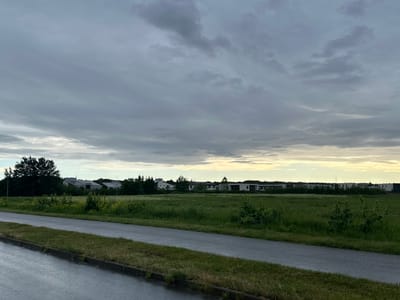The image depicts a cloudy sky over a rural landscape in Harkujärve, Estonia. In the foreground, a wet asphalt road runs diagonally from the bottom left corner towards the right. To the right of the road is a strip of grass, and then a large field of green vegetation. In the middle ground, a line of trees separates the field from a cluster of modern-looking white houses with dark roofs. The background consists of a dense line of dark green trees. The overall lighting suggests it is either early morning or late evening, with the clouds obscuring direct sunlight. No people or animals are visible in the scene.