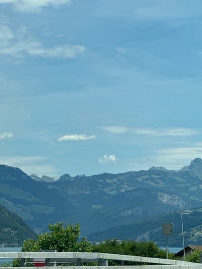 The image shows a panoramic view of a mountainous landscape under a bright, partly cloudy sky. In the foreground, a white metal guardrail runs horizontally across the frame. Behind the guardrail, lush green trees and foliage are visible. To the left, a glimpse of a body of water, likely a lake, can be seen. Further in the background, layered mountain ranges dominate the scene, with their peaks and slopes rendered in various shades of green and blue due to distance and atmospheric haze. Small white clouds are scattered across the vast blue sky. The location is specified as Buochs, Switzerland.