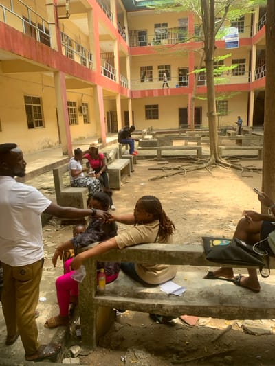 This image displays an unpaved outdoor courtyard surrounded by a multi-story building, identified as being in Calabar, Nigeria. The building has light yellow-painted walls, red trim on its structural elements and railings, and multiple accessible balconies or walkways on at least three visible floors. Numerous windows and doors are visible on the building's facades.
Approximately ten individuals are present in the scene. In the immediate foreground on the left, an adult male with a beard, wearing a white short-sleeved shirt, khaki trousers, and brown sandals, stands facing a group of seated individuals. He has his left arm extended, touching the upper arm of a female subject. This female is seated on a grey concrete bench, wearing a dark patterned top and bright pink leggings, holding an infant dressed in yellow. Next to her, another female in a tan short-sleeved top with braided hair leans against the bench's backrest, looking towards the interaction. Further down the same bench, a third female in a white top and patterned skirt is seated. To the left of this group, another female in a pink top and tan cap is seated on a separate concrete bench.
In the midground, a male subject in a dark jacket and red shoes is seated on the back of a concrete bench, facing away from the camera, appearing to interact with a handheld device. On the far right foreground, partially visible, are the legs of an individual wearing dark shorts and black sandals, next to a black shoulder bag and a hand holding a smartphone. In the background, on the second-floor balcony, a person walks from left to right. On the first-floor balcony to the far left, another person stands. On the ground level, far right, a male in a light blue shirt stands by a concrete bench, also looking at a handheld device.
The courtyard ground consists of reddish-brown dirt/sand, uneven and scattered with small debris, including a plastic bottle and papers. A prominent tree with exposed roots stands centrally in the courtyard. Multiple grey concrete benches are arranged throughout the space. Strong sunlight indicates a daytime setting, casting distinct shadows.