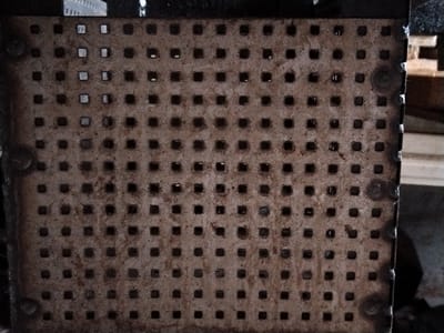 This media file shows a close-up, high-angle view of a weathered, perforated metal surface, likely a grate or a panel with numerous square holes. The metal is a rusty brown color, with signs of wear and tear, suggesting it is an outdoor object. The perforations are arranged in a regular grid pattern. Some of the holes appear to be darker than others, indicating shadows or debris within them. There are also some lighter patches, possibly due to reflections or cleaner areas of the metal. The texture of the metal is somewhat rough and uneven.

In the background, there are out-of-focus elements that suggest an industrial or storage setting. A dark, rectangular shape dominates the top edge of the image, and to the right, there are glimpses of lighter-colored structures, possibly wooden beams or crates, and a white object that might be part of a building or equipment. The overall lighting appears to be dim, suggesting it might be indoors or an overcast day. There is no visible text in the image. The focus is entirely on the texture and pattern of the perforated metal. The scene is devoid of any people or animals. The setting implies a functional, utilitarian object rather than a decorative one. The location context of Pargolovo, Russia, does not provide specific visual cues within the image itself, but it informs the potential context of such an object.