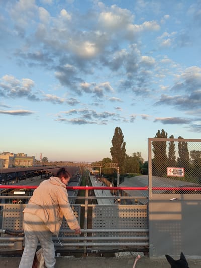 The image depicts a view from an elevated pedestrian walkway overlooking a railway station in Ramenskoye, Russia. In the foreground, a woman with reddish-brown hair pulled back is leaning over the railing, wearing a light-colored jacket and grey sweatpants. She appears to be looking down at the railway tracks below. The walkway itself is constructed of metal railings and concrete.

Beyond the walkway, the railway station is visible with multiple tracks, platforms, and some train cars. Buildings line the far side of the station. In the mid-ground, tall, slender trees stand alongside the railway infrastructure. A chain-link fence with a red warning sign that reads "ОСТОРОЖНО! ЭЛЕКТРИЧЕСКОЕ НАПРЯЖЕНИЕ" (Caution! Electric Voltage) is prominent on the right side of the frame.

The sky above is a mix of blue with scattered white and grey clouds, illuminated by what appears to be the golden hour light of late afternoon or early evening. The overall impression is of a typical urban scene at a transportation hub during a period of pleasant weather. The presence of the warning sign indicates that the area is a functional railway station with electrical hazards.