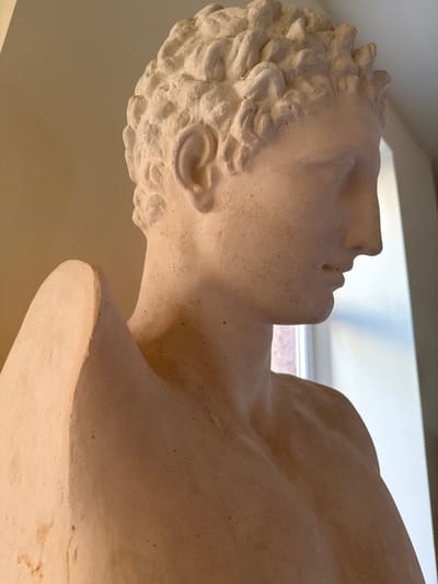 A light-colored, possibly plaster or cast stone, male bust is depicted in a three-quarter profile, facing right, within an indoor setting in Talin, Armenia. The upper torso, neck, and head of the figure are visible. Features include short, curly hair, a distinctly sculpted right ear, a prominent straight nose, and undetailed eye sockets. The mouth is slightly open. The material is off-white to cream with a matte, textured finish and scattered dark flecks. A sculpted element, appearing wing-like or as drapery, originates from the figure's upper left back/shoulder area, extending upwards. The background on the left consists of a plain, light-toned wall. The background on the right reveals a vertical window providing ambient light. No individuals or actions are present.
