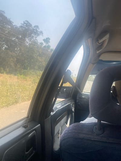 The scene depicts a view from the interior of a vehicle traveling on an unpaved road in Unguwar Gaya, Nigeria, under bright, hazy daylight conditions. The perspective is from a rear passenger seat, looking forward and towards the left.
Inside the vehicle, a dark-colored door panel with a visible door lock pin is present on the left. The vehicle's left A-pillar displays a seatbelt mechanism. On the right side of the frame, a dark fabric headrest, possibly denim, from a front seat is visible, partially obscuring the view. The back of a person's closely shaven or bald head is discernible above this headrest, indicating a front passenger or driver. A damaged, light-colored overhead grab handle is mounted on the vehicle's interior roofline to the right. Parts of the dashboard, steering column, and control panel are visible through the front passenger window.
The exterior environment consists of an unpaved or dirt road immediately adjacent to the vehicle on the left, flanked by dry, light-green and yellowish grasses and foliage. Beyond this vegetation, a dense line of green trees extends into the background. Multiple power lines run horizontally across the upper portion of the tree canopy. The foreground also shows more unpaved road and distant vegetation under a hazy, light-blue sky.