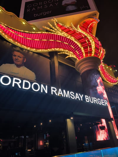 A nighttime, low-angle exterior shot captures the illuminated facade of Gordon Ramsay Burger, a restaurant located in Paradise, Nevada, United States. The restaurant's signage is prominently displayed, with large white neon letters spelling out "GORDON RAMSAY BURGER." Above this, a billboard features a black and white portrait of Gordon Ramsay in a chef's uniform, with his signature in script to the right. The restaurant is adorned with a large, elaborate structure resembling a flame, outlined with vibrant pink and yellow neon lights. This decorative element extends over the entrance. The overall scene is dark, with the bright lights of the signage and decorative features creating strong contrasts. The environment suggests a commercial or entertainment district, typical of Las Vegas.