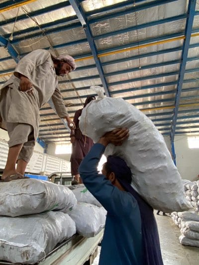 In an industrial warehouse in Shurandam, Afghanistan, three men are loading large, sack-like bags onto a truck. One man, wearing a light-colored robe and a head covering, stands on a stack of bags on the back of the truck and appears to be directing the loading process, holding a small white object in his hand. Another man, dressed in a blue top and a long dark skirt, hoists a large white bag onto his shoulder. A third man, wearing brown clothes, stands nearby. The warehouse has a corrugated metal roof with visible structural supports. Bags of similar size and shape are stacked to the right of the truck.