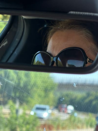 The image is a close-up shot of a person's reflection in a car's rearview mirror, taken from inside the vehicle. The reflection shows the upper part of the person's face, wearing dark-rimmed sunglasses, with their blonde hair visible. The background reflected in the mirror shows greenery and a hint of a blue sky. The foreground of the image, seen through the car's windshield, is blurry and shows more greenery, with two cars visible in the distance. One car appears to be silver with its headlights on, and another is red. There are also some smudges and streaks on the windshield. The scene appears to be an outdoor environment, likely on a road. The provided metadata indicates the location as Itzig, Luxembourg.