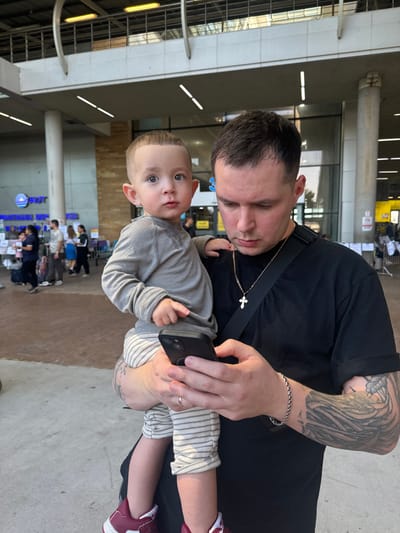 A man in a black shirt holds a baby in his arms while looking at a smartphone. The man has tattoos on his left arm and wears a silver bracelet. The baby wears a gray shirt, striped pants, and maroon shoes. They are standing in an airport. People and luggage are visible in the background.
