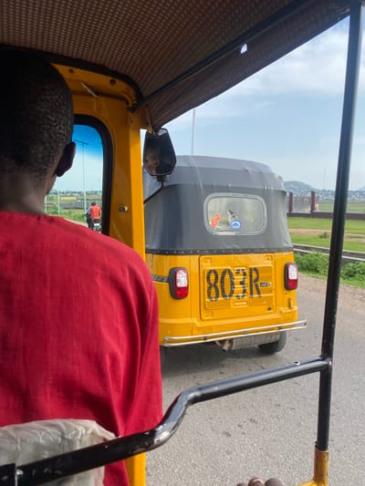 The image captures a perspective from within a yellow auto-rickshaw, also known as a "keke." The main subject in the foreground is the back of a person's head, with short, dark, textured hair. They are wearing a bright red garment that covers most of their upper body, suggesting they are a passenger. The interior of the keke is visible, with a patterned brown fabric lining the roof.

In the middle ground, another yellow auto-rickshaw is prominently featured, traveling ahead of the viewer's vehicle. This keke has a dark grey canopy and a bright yellow rear panel. Clearly visible on the rear panel is text which reads "803R" in large, bold, black lettering. There are also two red taillights, one on each side of the rear panel, and a silver exhaust pipe is visible on the left side of the keke. A circular blue and white sticker is affixed to the rear window of this keke.

The scene appears to be a roadside or street, with asphalt visible under the vehicles. In the background, there is a fence and some greenery, with hints of buildings or structures beyond, suggesting an urban or semi-urban environment. The sky is overcast with patches of blue, indicating daytime, possibly late morning or early afternoon. The overall impression is that of everyday travel or transit in Zanzi, Nigeria, characterized by the common mode of transportation and the bustling roadside environment.