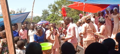 The image depicts an outdoor gathering, likely in a residential area within the city of Kalemie, Democratic Republic of the Congo. A large group of people of varying ages are present, some standing and others positioned behind a makeshift stage or platform.
Two men in the foreground, dressed in light pink outfits, appear to be speaking into microphones. Near them, a bright green bucket sits atop a woven basket. To the right of the speakers, a group of women in matching light pink outfits and head coverings seem to be a choir or musical ensemble. One woman is holding and playing a cymbal.
Other individuals in the crowd are wearing a variety of clothing, including shirts with text and patterned fabrics. Some are looking towards the speakers, suggesting they are listening to the presentation. Overhead, there are temporary coverings of blue and red fabric, possibly providing shade from the sun. The background shows trees and vegetation, with a hint of hills in the distance.