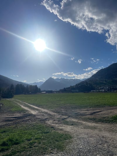 The image presents a landscape scene in Sterzing-Vipiteno, Italy, under a bright sun. The sky is blue with scattered white clouds. The sun's glare dominates the left side of the image. Mountains are visible in the background, partially obscured by clouds. In the foreground, there's a field of green grass. Adjacent to the grass, a dirt path or road is visible, indicating some level of traffic. The image appears to be taken during daylight hours.