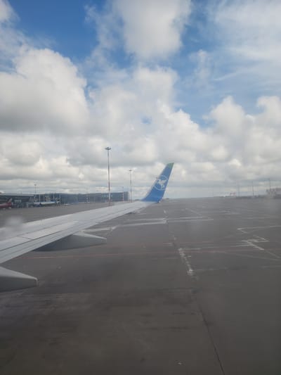 The image shows a view from an airplane window, likely at an airport in Bol'shie Kabany, Russia. The left side of the frame is dominated by a bright blue sky with scattered white clouds. In the lower part of this section, the blue tail fin of the airplane is visible, emblazoned with a light-colored emblem. The right side of the image displays a section of the airplane wing, painted white, with visible flaps and components. Beyond the wing, the airport tarmac stretches into the distance, appearing as a dark gray surface with faint lines. Various airport structures, including buildings and light poles, are visible along the horizon line. The overall scene conveys the feeling of preparing for takeoff or shortly after landing.