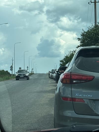 The image depicts a traffic jam on a road in Kachimir, Russia, under a cloudy sky. A line of cars is stationary on the asphalt road, with trees and utility poles lining the sides. In the foreground, the rear of a grey SUV is visible, with the "TUCSON" logo clearly displayed on the tailgate. Further down the road, a black Mercedes-Benz is approaching the traffic. Several streetlights are also visible along the road, indicating it's likely a main thoroughfare. The overall scene suggests a typical day in a city where traffic congestion is common.