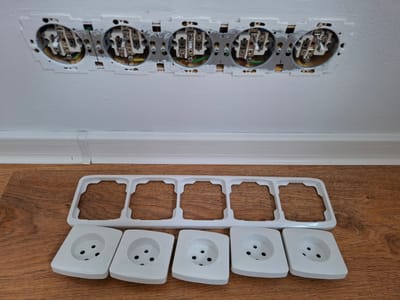 This media file shows a partially disassembled electrical outlet system. In the background, mounted on a white wall, are the internal components of five electrical outlets. These components appear to be in the process of being installed or removed, with wires and terminals visible.
In the foreground, resting on a wooden floor, are the external parts of the outlet system. There is a white plastic frame that would hold multiple outlets, and five individual white outlet covers. Each outlet cover has the standard three circular holes for plugs.
The scene appears to be an indoor setting, likely a room under renovation or maintenance. The lighting suggests it is daytime, but there are no specific indicators of the time of day or weather. The location context provided is Bratislava, Slovakia, which is a factual piece of information about the image. There is no visible text in the image. The overall impression is one of a technical task involving electrical work.
