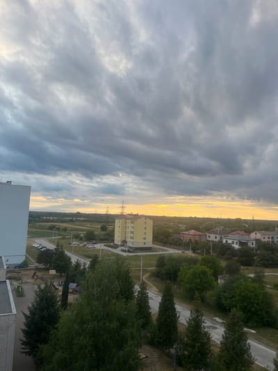 This is a vertical, medium-distance, outdoor, full-shot photograph of a suburban landscape in Dobrotvir, Ukraine, during twilight. The sky is filled with dramatic, heavy grey clouds, with a bright yellow band of sunlight visible on the horizon, suggesting the sun is setting or has just risen. In the foreground, to the left, is the side of a tall, modern, white-and-grey apartment building. To the right and slightly further back, a smaller, bright yellow apartment building with visible windows and balconies stands. A small, undeveloped playground with colorful slides and swings is visible between the two buildings. Beyond the apartment buildings, there is a road curving through a grassy area with scattered trees and smaller residential houses with red and white roofs. In the distance, there are power lines and transmission towers, and the horizon is lined with trees. The ground appears to be a mix of pavement, grass, and dirt. There are no people visible in the frame.