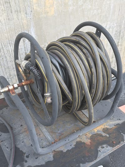 An outdoor, medium-shot, eye-level photograph shows a heavy-duty, black rubber hose coiled onto a metal hose reel. The hose has two parallel yellow stripes running along its length. The reel is mounted on a wheeled cart, also made of dark metal. The cart appears to be positioned outdoors, on a grey, weathered surface with visible rust spots and water stains. In the background, a rough, textured concrete wall is visible. A rusted metal coupling is attached to the end of the hose, with another silver metal fitting connected to it. The overall scene suggests an industrial or maintenance setting in Murfreesboro, United States of America.