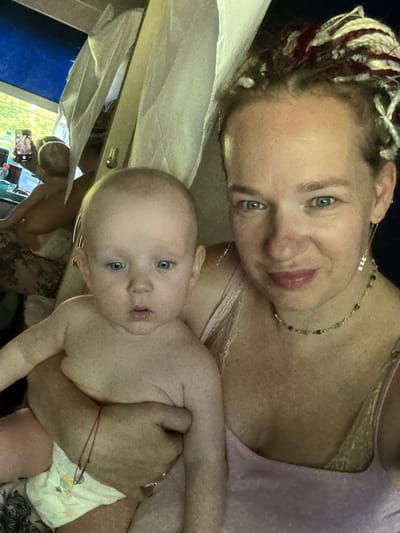 A woman with dreadlocks and blue eyes, wearing a pink top and a beaded necklace, holds a baby with blue eyes. The baby is shirtless and wearing a diaper. In the background, another baby is visible. A smartphone is being held up to take a photo, showing a screen with an image of the two babies. The setting appears to be indoors, possibly a home, with a view of greenery through a window. The image is a selfie taken by the woman. The location is specified as Gal'chino, Russia.