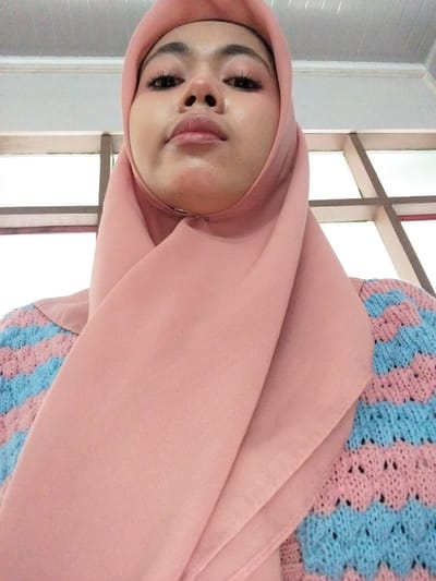 A low-angle, close-up, selfie-style photograph shows a young woman wearing a pink hijab and a knitted sweater with horizontal stripes of pink and light blue. She is looking upwards towards the camera with her eyes open and a slight smile. Her makeup includes pink eyeshadow, mascara, and lip gloss. The background consists of a white ceiling with horizontal paneling and a window frame with dark brown or black mullions. The lighting suggests an indoor setting, possibly a room with natural light coming from the window. The image is taken from the chest up, focusing on the woman's face and upper body. No specific location in Tibojong, Indonesia, is identifiable from the image itself.