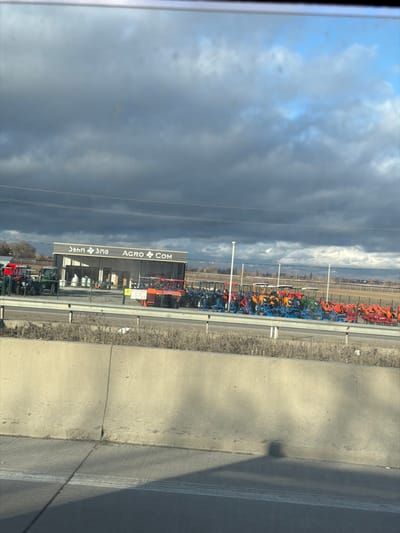 The image depicts an outdoor agricultural equipment sales lot under a heavily clouded sky in Akhalsopeli, Georgia, viewed from a moving vehicle on a highway. In the foreground, a concrete highway barrier separates the observer's lane from the adjacent area. Beyond the barrier, a patch of dry, light-colored vegetation borders a property. Centrally positioned behind this vegetation is a grey, single-story commercial building featuring a sign that reads "ჯიბი ჯმშ" in Georgian script and "AGRO COM" in English, indicating an agricultural business. The area surrounding and extending to the right of the building is an open display lot filled with numerous new agricultural machines and implements. These include green tractors, blue cultivators or plows, orange loaders, and various other colorful farm machinery arranged in rows. Multiple white utility poles are spaced across the display lot. In the distant background, a flat landscape with a faint line of trees or low hills is visible beneath a sky dominated by dark grey, dense clouds, with hints of lighter blue sky peeking through towards the upper right. No individuals, specific actions, or interactions are discernible in the scene.