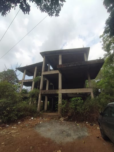 An unfinished, multi-story concrete building stands in an overgrown lot in Marna, India. The structure consists of support pillars and floor slabs, without walls or other finishing. Foliage partially obscures the building. A dark-colored vehicle is visible at the edge of the frame. The ground is covered in dirt and debris. Overcast sky.
