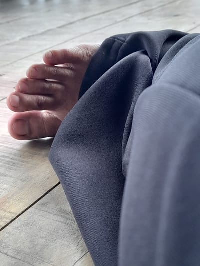 The image depicts a close-up view of a bare human left foot and the lower portion of a leg, clothed in dark grey fabric, resting on a light-colored wooden plank floor. This scene is located in Penanggungan, Indonesia. The foot displays five toes with visible, unpolished toenails. The fabric on the leg, likely from trousers or sweatpants, exhibits creases and textile texture. The wooden floor comprises multiple planks with visible grain patterns and seams. The subject is stationary and at rest; no other individuals or objects are present within the frame. Natural illumination highlights the textures of the skin, fabric, and wood.