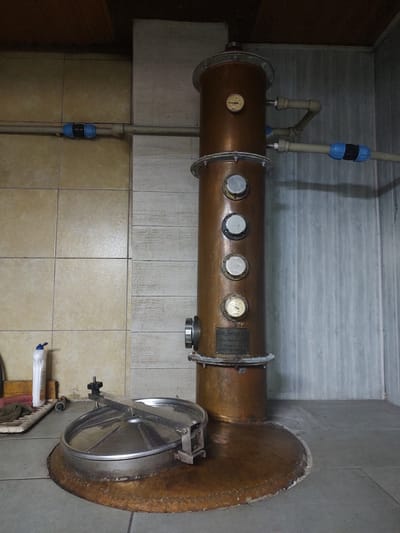 The image displays a large, copper cylindrical apparatus with various gauges and fittings, likely a distillation still. It is situated in what appears to be a workshop or utility area, with tiled walls and floors. Several pipes are connected to the apparatus, suggesting it is part of a functional system. A large, circular metal hatch with a locking mechanism is attached to one end of the cylinder.
The overall setting is industrial or functional, with plain walls and tiled surfaces. There are no people visible in the image. The lighting appears to be artificial, as there are no natural light sources evident. The focus is entirely on the distillation equipment.
A visible label on the copper cylinder contains text that reads: "ICA - ДECHIL", "Техн. Бренд.", "1- Состав", and "GSM - 0.035 m". The remaining text on the label is illegible.
The image is taken from a slightly elevated angle, looking down at the apparatus. The copper has a patina, indicating age or exposure. The scene appears to be indoors, with no indication of the time of day or weather conditions. The context provided of Kochmar, Bulgaria, suggests a possible artisanal production setting.