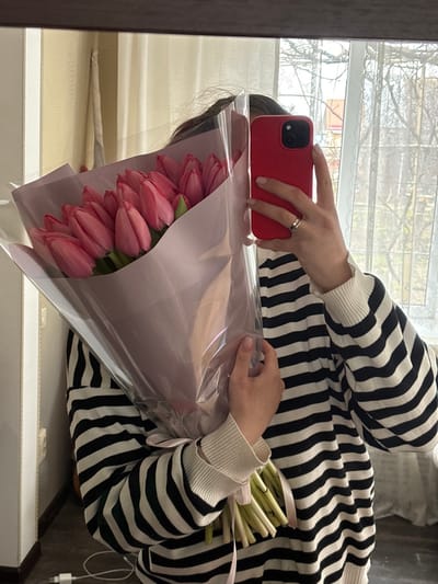 A mirror selfie captures an individual from the chest up, reflecting in a dark-framed mirror indoors. The person wears a black and white horizontally striped long-sleeved top with white ribbed cuffs. Their face is largely obscured by dark hair and a large bouquet of pink tulips held with the left arm. Approximately 20-25 closed pink tulips, wrapped in light pink paper and clear cellophane, are visible. The individual's right hand holds a red-cased smartphone (likely an iPhone) at an angle, indicating the capture of the reflection. A simple band-style ring is visible on the ring finger of the right hand. Both visible hands have neatly manicured nails with clear or light polish. The reflected background reveals a light-colored wall or curtain on the left and a window offering a view of bare tree branches and a distant red-roofed building, typical of a residential area. An electrical outlet with a white cord and adapter is visible on the lower left wall. The image provides no specific identifiable landmarks to confirm the location as Slavyansk-na-Kubani, Russia.