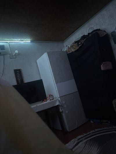 The image shows an interior scene, possibly a residential room, in Tsar Kaloyan, Bulgaria. The room is dimly lit, with artificial lighting from an LED strip along the ceiling and a visible light bulb. The primary object in the scene is a tall, white refrigerator, which appears to be positioned at an angle. To the left of the refrigerator, a black television is visible, and above it hangs a framed picture. On a surface in front of the TV and refrigerator, a decorative object with a patterned surface, possibly a ceramic figure or a lamp, is present, with a small red light emanating from it. A portion of a person's arm or leg, out of focus and in the foreground, suggests the presence of an individual. In the background, a large, dark object, possibly a wardrobe or bed with coverings, dominates the right side of the frame. A pink pouch or bag rests on this dark object. The walls have a textured wallpaper, and the ceiling is made of wood-like panels. A rug with a striped pattern and fringes is visible on the floor. The overall impression is of a cluttered and somewhat unkempt living space.