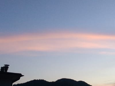 The image captures a serene twilight scene over Valle Castellana, Italy. A silhouette of a house with a chimney stands against the fading light on the left side, while the dark outline of a forested hill stretches across the lower right portion of the frame. The sky is a gradient of soft blue and pink hues, with a delicate band of pink clouds stretched horizontally across the middle. The overall atmosphere is tranquil and picturesque, indicative of a peaceful evening.