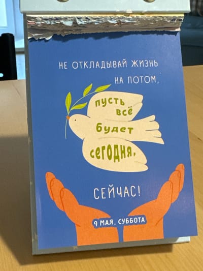 Here's a forensic description of the image:
The image shows a desk calendar, open to a page with a blue background. The text on the page is written in Russian, and the illustration includes a white dove with an olive branch, surrounded by orange hands. The text reads "НЕ ОТКЛАДЫВАЙ ЖИЗНЬ НА ПОТОМ," which translates to "Don't put off life until later." The next lines, "пусть всё будет сегодня," translates to "let everything be today," and "СЕЙЧАС!" means "NOW!" Below the hands it reads, "9 МАЯ, СУББОТА," which translates to "May 9th, Saturday". The location of the image is likely an indoor space. There is no indication of Люк or Russia.