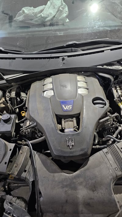 An overhead shot shows the engine bay of a car, with a prominent V6 engine cover bearing the Maserati trident logo. The engine is covered in a layer of dust. To the top of the image, a crumpled white cloth is visible on the windshield. The car is parked outdoors in Ataşehir, Turkey, under what appears to be daylight. The scene suggests the vehicle is likely parked on a street or in a parking lot.