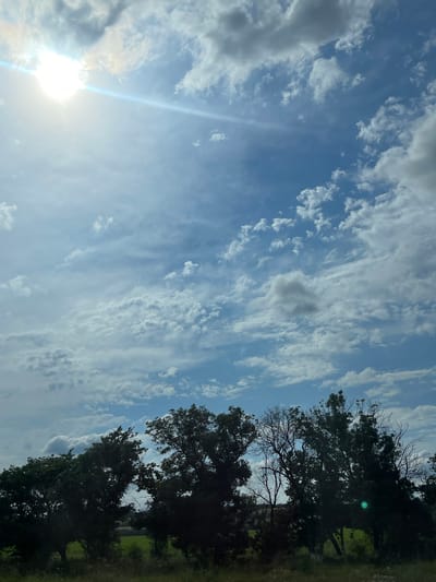 The image depicts a bright, partly cloudy sky over a landscape with trees and grass. The sun is visible on the left side of the frame, casting a lens flare across the sky. The clouds are a mix of white and gray, with some appearing wispy and others more substantial. The trees are dark green and silhouetted against the sky, suggesting they are in the foreground. The grass appears to be a light green, indicating it might be a field or park. The overall impression is one of a pleasant day outdoors. The time of day appears to be daytime, likely late morning or early afternoon, given the brightness of the sun. The weather is partly cloudy, with a mix of sunshine and cloud cover. There is no text visible in the image. The scene is a natural landscape, likely in a rural or suburban area.