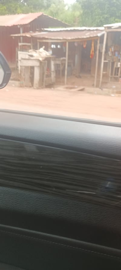 The image, likely taken from inside a moving vehicle, shows roadside commercial establishments in Ozalla, Nigeria. The establishments appear to be small shops or stalls constructed with wood and corrugated metal roofing. The road is unpaved, showing a reddish-brown color. There are no discernible people visible. The interior of the car, including the dashboard, is visible in the foreground.
