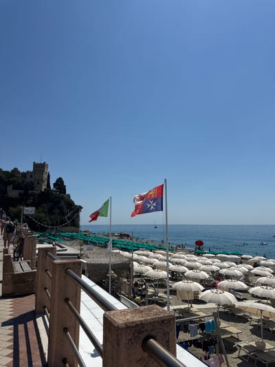 The image depicts a bright, sunny day at a beach in Finale Ligure, Italy. The scene is dominated by rows of white umbrellas and lounge chairs set up on a pebble beach, indicating a popular spot for sunbathing and relaxation. The calm, blue sea stretches towards the horizon under a clear sky.

Several flags are visible, adding a touch of national and regional pride to the scene. A prominent flag, likely representing a maritime republic or a historical entity, features red and white sections with a white cross on a blue background. Another flag is the Italian tricolor, green, white, and red, flying alongside it.

In the background, a picturesque castle or fort is perched on a lush, green hillside, adding a historical and scenic element to the coastal view. People are scattered across the beach and in the water, suggesting a leisurely day. The time of day appears to be midday or early afternoon, given the strong sunlight and shadows. The overall mood is one of a pleasant, typical summer day by the Italian Riviera. A sign with text is visible on the hillside, but the lettering is too small to be legible.