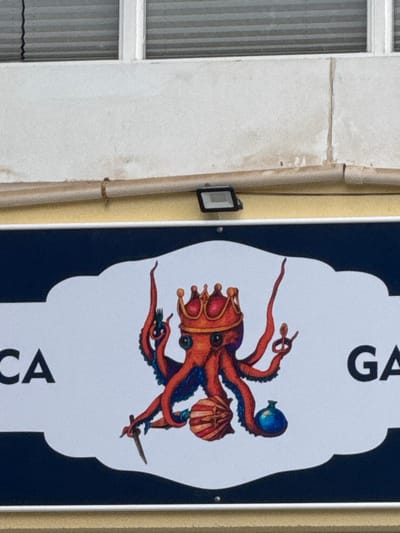 The image displays a portion of a building facade in Barriada De Villalba, Spain, primarily featuring a sign. The sign is white with an ornate, cloud-like top and bottom edge, bordered in dark blue. Visible text fragments on the sign include "CA" in black capital letters on the left and "GA" on the right. Centered on the sign is an illustration of an orange-red octopus with two prominent eyes, wearing a gold and red crown. The octopus uses its tentacles to grasp various items: a green-handled fork is held in its upper left tentacle, a gold ring in its upper right, a silver knife in its lower left, a red and white striped scallop shell centrally below it, and a blue bulbous container in its lower right tentacle.
Above the sign, a section of yellow-painted wall is visible, on which a black rectangular floodlight is mounted. A light brown conduit or pipe runs horizontally across this yellow wall, secured by several brown ties. Above the conduit, the wall transitions to a white, textured surface with vertical cracks or seams and areas of discoloration. The top edge of the image reveals a window with a white frame and horizontal venetian blinds visible inside. No individuals are present in the scene.
