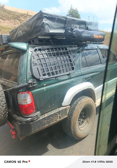 The image displays the rear three-quarter left view of a dark green, mud-splattered sport utility vehicle, consistent with a Toyota 4Runner model, in motion on a paved road under a bright, partly cloudy sky. The vehicle is outfitted with a black roof rack supporting a large, black, soft-sided rooftop tent or storage bag labeled "ARK ROMA," and a smaller black cylindrical bag marked "SAN HIMA" in orange lettering. A black, grid-patterned security or cargo panel, bearing "ART" branding, is affixed over the rear left side window. An aftermarket black bumper features a spare tire mounted on the rear left and a red recovery shackle. Large, muddy off-road tires on black rims are visible, exhibiting motion blur. Light-colored aftermarket fender flares protect the wheel wells. The background shows a sloping dirt embankment or hillside on the left and minimal vegetation. No individuals are distinctly visible within the vehicle. The scene is situated in Lari/Kirenya ward, Kenya.
