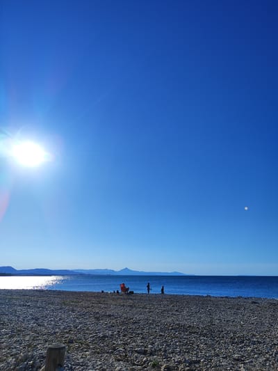 The image depicts a bright, sunny day at a pebble beach in Els Poblets, Spain. The sun is high in the clear blue sky, casting a bright glare on the tranquil, deep blue Mediterranean Sea. In the distance, a range of mountains forms a hazy silhouette against the horizon.
On the beach, a few people are visible. Two individuals stand near the water's edge, one appearing to be holding a fishing net. Further to the right, a single orange beach chair is set up, with what appears to be some belongings beside it. The beach itself is covered in smooth, gray pebbles.
The scene suggests a peaceful, recreational day at the coast, with the natural beauty of the landscape being prominent. The overall impression is one of a serene seaside environment under a clear sky.