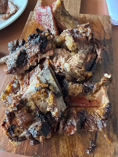 A close-up shot reveals a pile of cooked meat, likely ribs, served on a wooden cutting board. The meat exhibits a charred exterior with visible bones interspersed throughout. The overall color palette is dominated by shades of brown, ranging from the dark, burnt edges to the lighter hues of the meat's interior. A glimpse of a white plate with more meat is visible in the lower left corner.