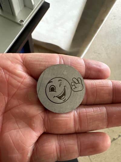 The image shows a person's palm holding a round, metallic object. The object has an engraved design of a face with an exaggerated expression of surprise or distress, with wide eyes and an open mouth. Above the face, there is a crudely drawn hand symbol with two fingers extended. The palm displays prominent lines and textures, indicating an adult hand. The background is blurred, but suggests an indoor setting with a light-colored surface and a darker object. The lighting appears to be natural, casting soft shadows. The overall impression is that the object is a token or a charm with a humorous or expressive design. There is no visible text in the image.