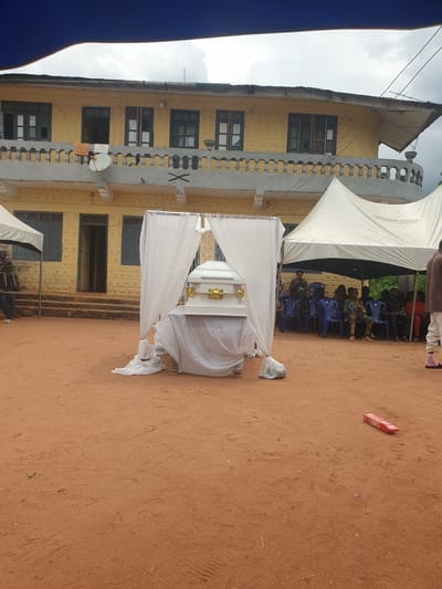 The image shows an outdoor scene in Nkwerre, Nigeria. A white coffin, draped in white cloth and adorned with a gold cross, stands under a white canopy. A two-story yellow building with dark-framed windows serves as a backdrop. Attendees sit in blue plastic chairs under a larger white tent in the background. The ground is red soil. A red and white package lies on the ground.