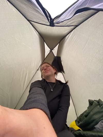The image shows a first-person perspective inside a tent. The tent's interior fabric is visible, with numerous water droplets indicating it is raining outside. A young woman is partially visible, lying down and looking upwards with her eyes closed and a slight smile, suggesting a sense of contentment or relaxation. She is wearing a black long-sleeved shirt and a silver pendant necklace. To her right, there are piles of camping gear, including a dark green sleeping bag and a yellow object, possibly another sleeping bag or a mat. The overall environment suggests a rainy day spent camping. The location is noted as Абзаново, Russia, though no visual cues in the image specifically confirm this location.