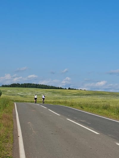 Two individuals are cycling on a paved road bordered by a vibrant green field under a clear blue sky with scattered white clouds. The road has distinct white lane markings. The field is lush, with some areas appearing lighter, possibly indicating wildflowers or a different type of grass. A line of trees forms the distant boundary of the field. The perspective suggests the photo was taken at eye level, with the road curving away into the distance. The lighting and clear sky indicate it is daytime, likely a sunny day. The overall scene evokes a sense of outdoor activity and peacefulness. The location is identified as Chumna, Russia.