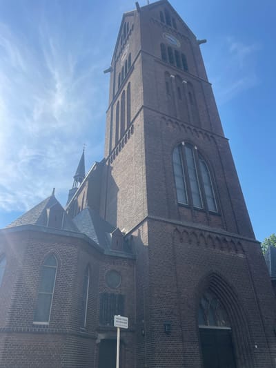 This is an outdoor, low-angle, eye-level shot of a church on a sunny day. The sky is bright blue with wispy white clouds. The church is made of dark red brick and has a tall, square tower. The tower has several arched windows and a clock face with a blue background and white hands. The church also has a sloped, dark-colored roof with several smaller spires. One spire has a cross at its peak. The building is illuminated by sunlight, casting shadows on some parts of the facade. A sign is visible on the right side of the church, featuring text in Dutch. Part of the text reads "verboden toegang voor onbevoegden." This translates to "no trespassing for unauthorized persons." The scene appears peaceful and serene. The bright sunlight and clear blue sky suggest it is daytime, likely late morning or early afternoon. The architectural style and the Dutch text on the sign indicate the location is in the Netherlands, specifically Venlo. No people are visible in the image. The focus is on the grand architecture of the church against the backdrop of a clear sky.